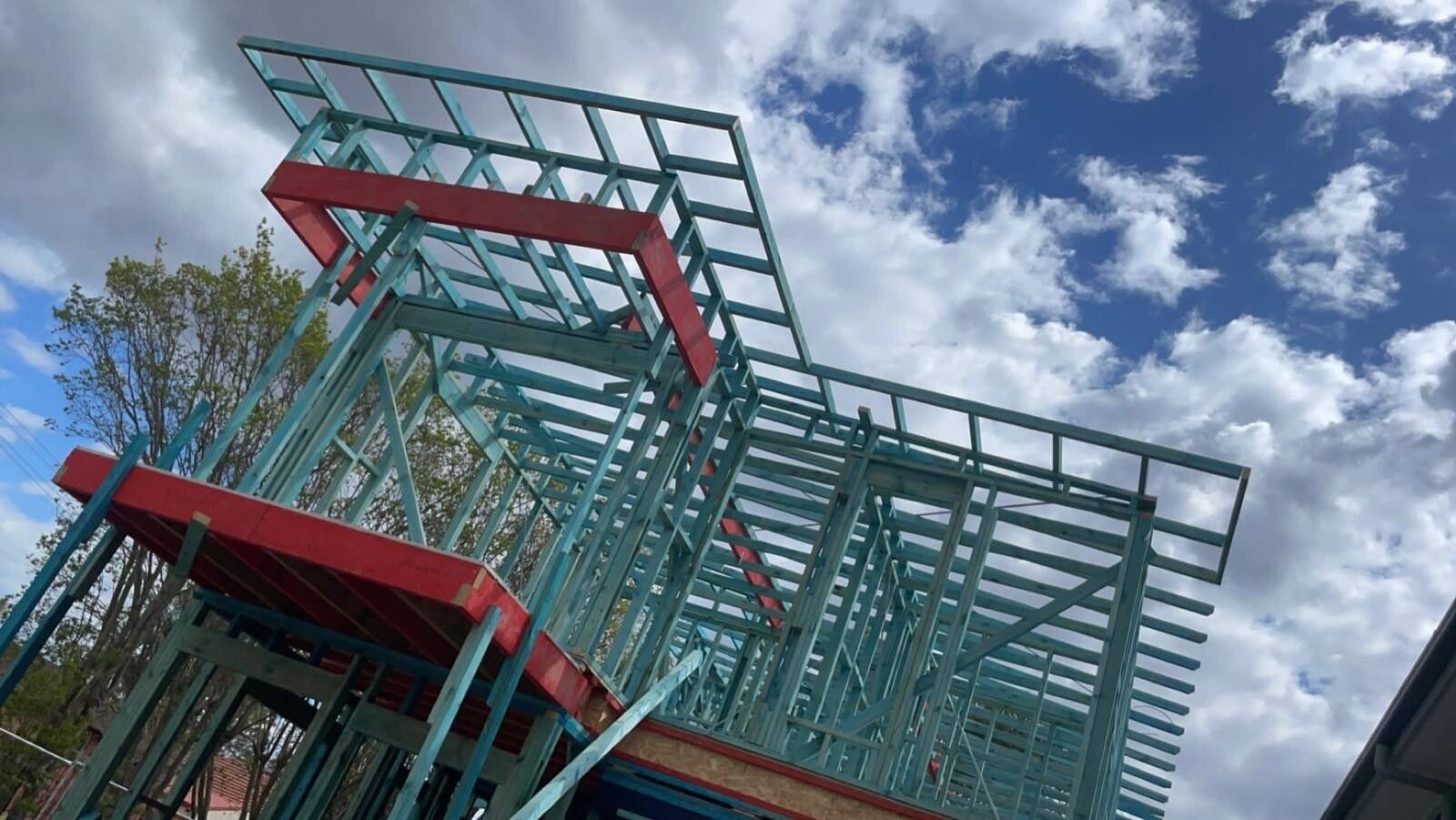 Low-angle view looking up at an upper floor timber frame: blue treated pine studs, top plates and joists with red LVL beams, structural flooring visible at floor level, against a blue sky with clouds